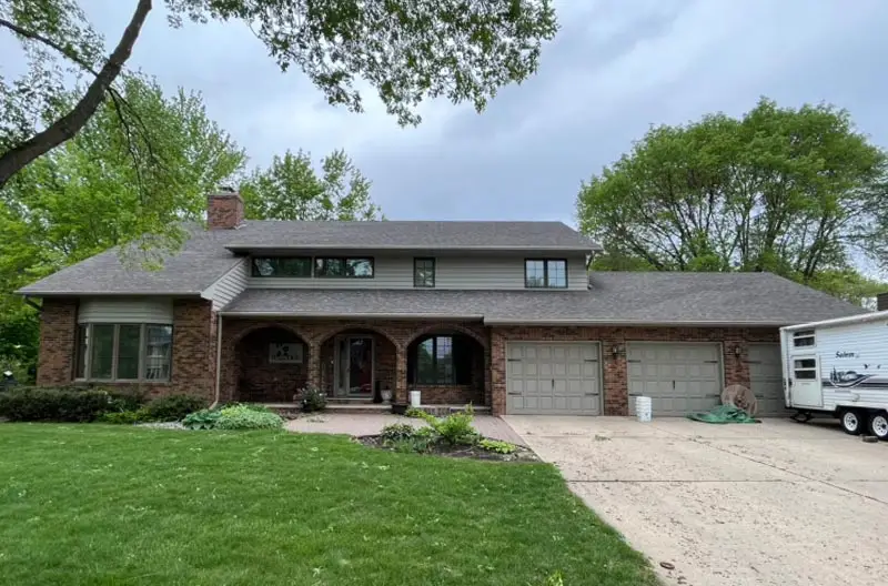 The 'before' photo of a two-story brick house in need of some exterior love. The faded siding, worn roof, and overgrown landscaping are ready for a refresh.
